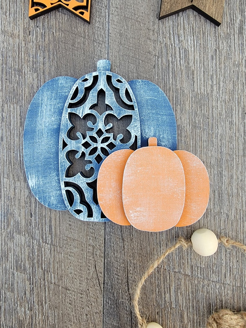 May include: Two wooden pumpkins, one blue with a cutout design and one orange, on a wood background.