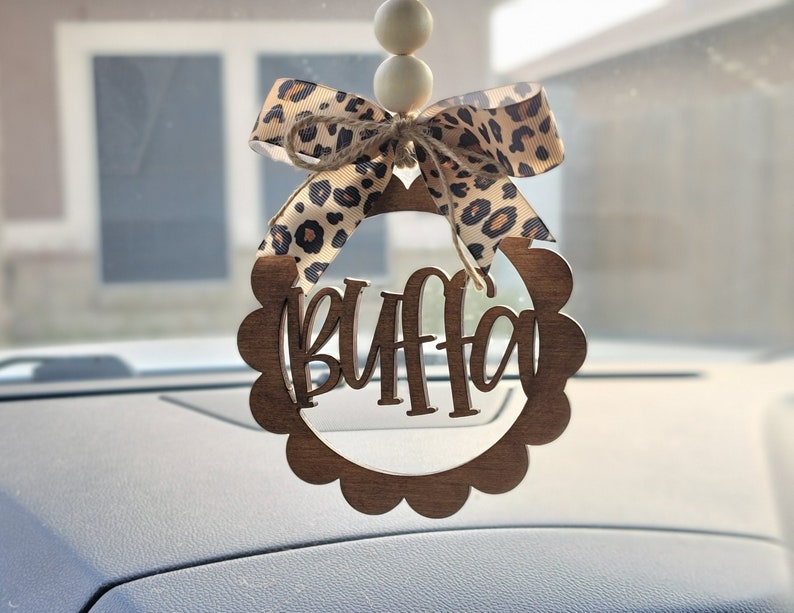 May include: A wooden car charm with the word "BUFFA" in a decorative font. The charm is decorated with a brown and white leopard print bow and white beads.