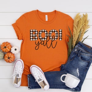 Puede incluir: Camiseta naranja con la frase "BOO! y'all" escrita en letras de cuadros negros y blancos.