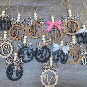 May include: A collection of 12 wooden ornaments with different letters and designs. Each ornament has a beaded string and a bow. The ornaments are in various colors, including black, brown, and white.