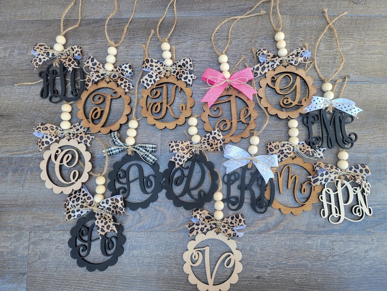 May include: A collection of 12 wooden ornaments with different letters and designs. Each ornament has a beaded string and a bow. The ornaments are in various colors, including black, brown, and white.