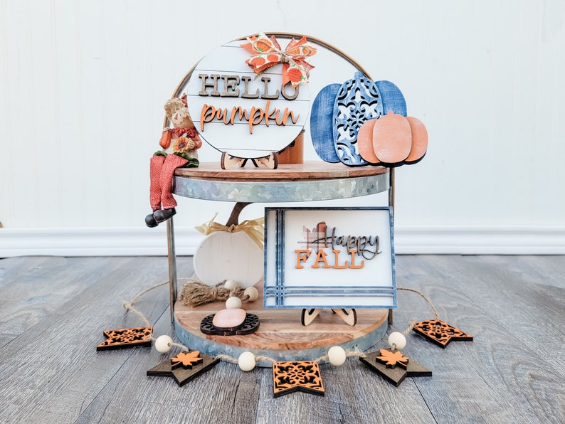 May include: A two-tiered metal stand decorated with fall-themed wooden signs. The top tier features a round sign that says "HELLO pumpkin" and a blue pumpkin with a smaller orange pumpkin. The bottom tier features a sign that says "Happy FALL" and a small white pumpkin. A wooden garland with orange and brown leaves hangs in front of the stand.