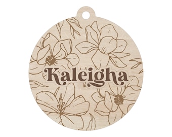 May include: A round wooden ornament with a floral design and the name "Kaleigha" in a brown, retro font. The ornament features a light wood grain background with brown floral outlines, and a small hole at the top for hanging.