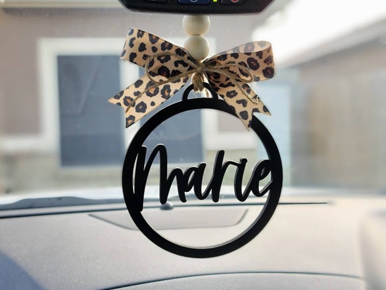 May include: Black and white car charm with the name "Marie" written in a cursive font. The charm is attached to a leopard print bow and a string of white beads.