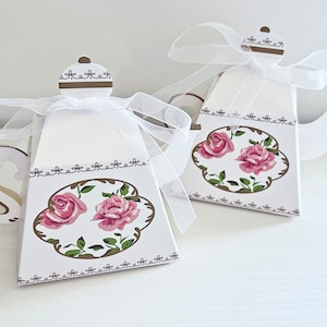 May include: Two white teapot-shaped gift boxes with pink roses and green leaves on the front. The boxes have brown trim and are tied with white ribbon.