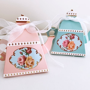 May include: Two decorative teapot shaped boxes with floral designs. The box on the left is pink with a white ribbon and the box on the right is blue with a white ribbon.