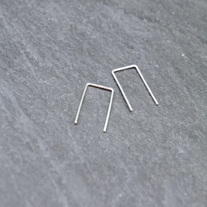 May include: Two minimalist silver-colored, U-shaped earrings. The earrings are small and simple, with a clean, modern design. They are set against a dark gray, textured background.