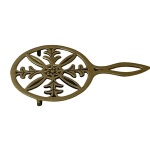 May include: A brass trivet with an oval shape and a handle. The trivet features a decorative openwork design with a central starburst and stylized leaf patterns. It has small feet to elevate it from the surface.