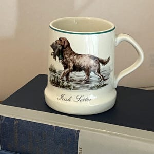 May include: A cream-coloured ceramic mug featuring a detailed illustration of a brown Irish Setter dog carrying a bird in its mouth. The mug has a green rim, a sturdy handle, and the text Irish Setter printed in black script near the base.