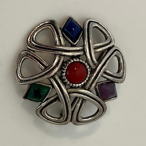 May include: Silver-toned brooch with a Celtic knot design. The brooch features a central red stone surrounded by four colored stones: blue, green, and purple. The brooch is a circular shape.