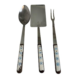 Puede incluir: Un juego de utensilios de cocina vintage que incluye una cuchara, una espátula y un tenedor. Las herramientas de acero inoxidable tienen mangos marrones oscuros con un diseño floral blanco y azul. El juego muestra signos de desgaste.