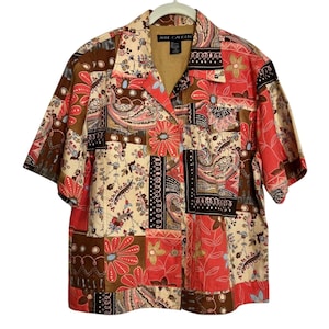 Puede incluir: Una camisa de patchwork de manga corta con cuello. La camisa presenta una variedad de estampados florales y paisley en tonos rojos, marrones y crema. La etiqueta dice "ANNE CALSON".