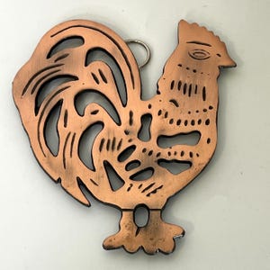 Puede incluir: Un adorno de pared de metal color cobre con forma de gallo, con intrincados detalles recortados en el cuerpo y las plumas de la cola. La superficie presenta líneas grabadas y una pequeña anilla metálica circular en la parte superior.