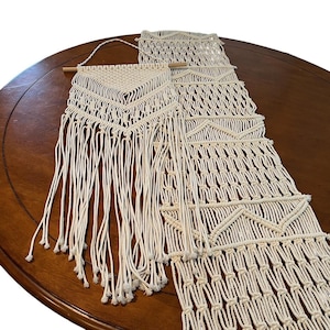 May include: Cream-colored macrame wall hanging with geometric patterns and fringe. The handmade textile features a wooden dowel for hanging and adds a bohemian touch to any space. The macrame design includes various knotting techniques.