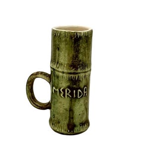 May include: A green ceramic mug with a bamboo-like design and a handle. The word "MERIDA" is written in white letters on the side of the mug. The mug has a light brown interior.