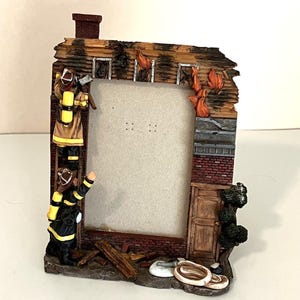 Puede incluir: Un marco de fotos rectangular diseñado como un edificio de ladrillo en llamas. Dos bomberos con equipo beige y negro aparecen en el lado izquierdo, uno en una escalera. El marco presenta llamas naranjas, una chimenea y una manguera enrollada.