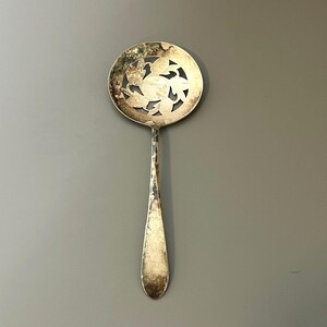 May include: A vintage silver-toned serving spoon with a round, perforated bowl featuring a floral design. The spoon has a long, slender handle and shows signs of age and wear, adding to its antique charm. The background is a solid gray.