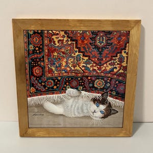 May include: A square framed print featuring a painting of a white cat with blue eyes lying on a light-colored floor. The cat is partially covered by a fringed rug with a detailed red, blue, and yellow pattern. The artist signature is visible.
