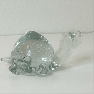 Puede incluir: Una figura de tortuga de cristal transparente con un caparazón redondeado lleno de pequeñas burbujas de aire. La tortuga tiene una cabeza, cuatro aletas y una cola corta. La figura se exhibe sobre una superficie blanca.
