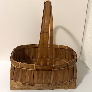 Puede incluir: Una cesta rectangular tejida con tiras de bambú marrón que presenta un asa alta y arqueada unida al centro de los lados largos. La cesta tiene una base plana y listones verticales reforzados con tiras de unión delgadas y más claras.