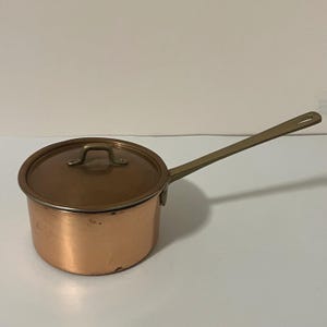 May include: A round copper saucepan with a matching lid and a long, straight brass handle. The pot features a polished metallic finish with visible signs of use, including minor surface variations and a sturdy, riveted handle attachment.