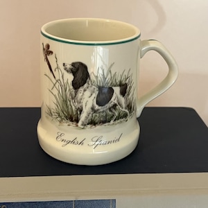 Può includere: Una tazza in ceramica color crema con un'illustrazione dettagliata di un English Spaniel bianco e nero in piedi nell'erba alta verde, con un piccolo uccello che vola sopra. Il testo English Spaniel è stampato in nero vicino alla base.