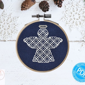 May include: A white angel cross stitch pattern on a dark blue background in a wooden embroidery hoop. The pattern is surrounded by white snowflakes and pine branches on a white wooden background. The text "INSTANT DOWNLOAD" and "CROSS STITCH PATTERN" are printed on a blue circle with the word "PDF" in white.