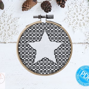 May include: A black and white cross stitch pattern of a star in a hoop. The hoop is made of wood and has a natural finish. The star is white and the background is black. The pattern is surrounded by white snowflakes and pine branches.