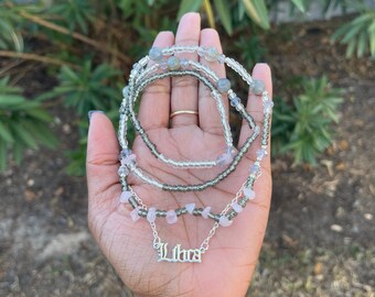 May include: A collection of beaded necklaces and bracelets. The bracelets and necklaces are made with clear, gray, and pink beads. A silver necklace with the word "Libra" is also included.