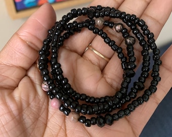 May include: A black beaded bracelet with pink and brown beads. The bracelet is wrapped around a hand.