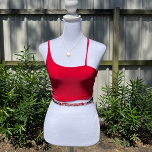 May include: A red one-shoulder crop top with a beaded belt. The top is on a white mannequin against a wooden fence background.