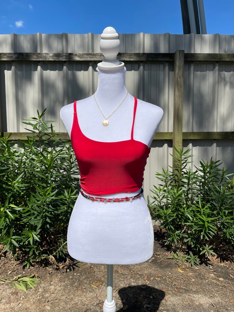 May include: A red one-shoulder crop top with a beaded belt. The top is on a white mannequin against a wooden fence background.