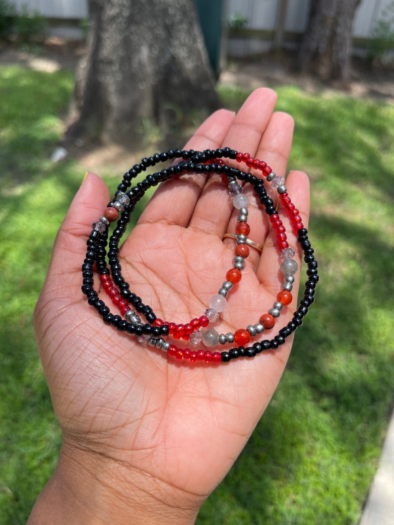 May include: A beaded waist chain with black, red, and clear beads. The chain has silver accents and is held in a hand.