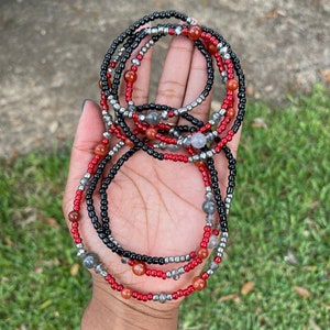 May include: A set of beaded waist beads in red, black, and silver. The beads are strung on a thin cord and are arranged in a layered pattern.