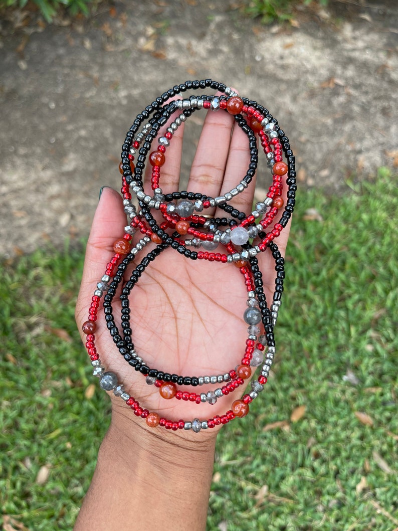 May include: A set of beaded waist beads in red, black, and silver. The beads are strung on a thin cord and are arranged in a layered pattern.