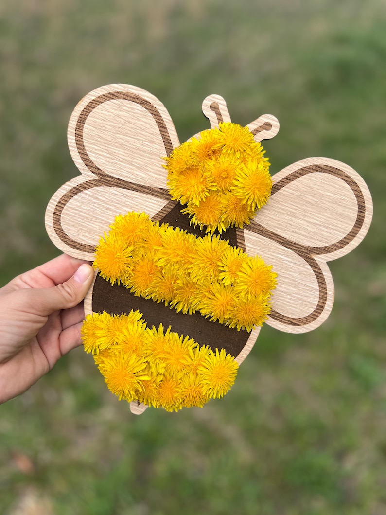 Puede incluir: Un recorte de abeja de madera decorado con flores de diente de le&oacute;n amarillas. La abeja tiene un cuerpo rayado de negro y marr&oacute;n.
