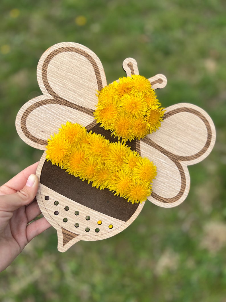 Puede incluir: Corte de abeja de madera con un cuerpo rayado marr&oacute;n y negro. La abeja est&aacute; decorada con flores de diente de le&oacute;n amarillas.