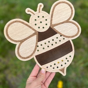 Puede incluir: Juguete de madera con forma de abeja con rayas marrones y blancas y una cabeza marr&oacute;n. La abeja tiene agujeros para enhebrar cuerda o hilo.