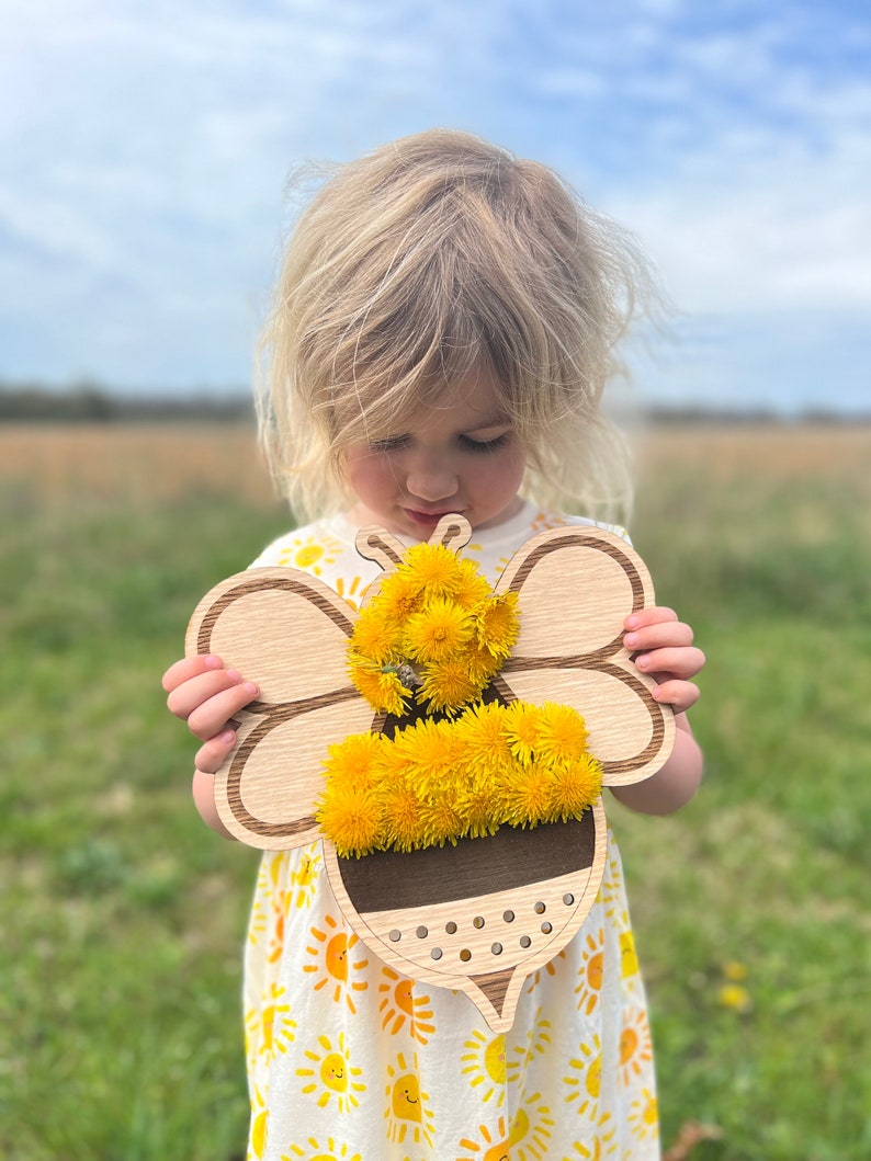 Puede incluir: Un ni&ntilde;o sostiene un recorte de abeja de madera con flores de diente de le&oacute;n amarillas adheridas. La abeja tiene una franja marr&oacute;n y un cuerpo texturizado. El ni&ntilde;o lleva un vestido blanco con soles amarillos impresos.
