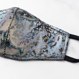 May include: A silver and black face mask with an iridescent pattern. The mask has a black elastic band.