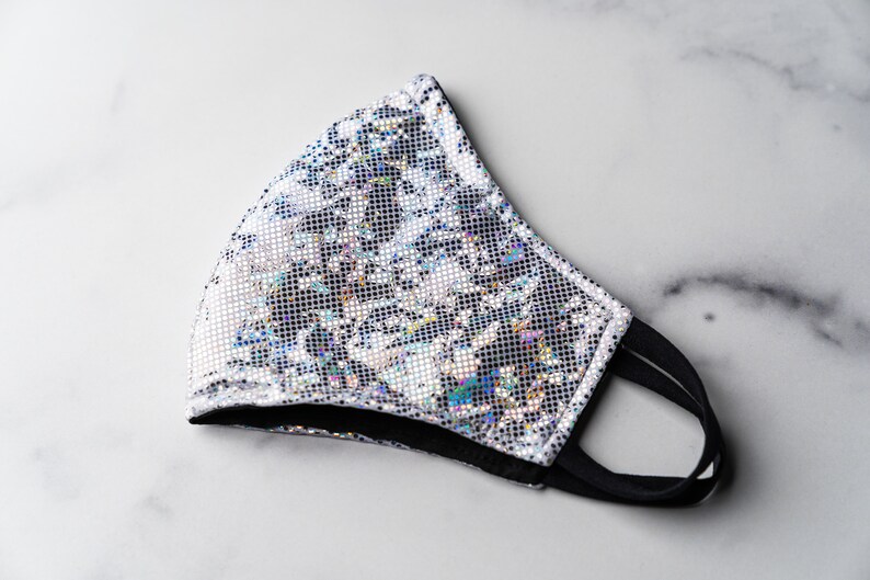 May include: A silver iridescent face mask with a black elastic band. The mask has a geometric pattern.