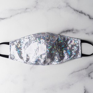 May include: A silver iridescent face mask with black elastic ear loops. The mask has a shimmery, holographic finish.