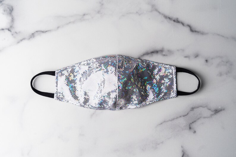 May include: A silver iridescent face mask with black elastic ear loops. The mask has a shimmery, holographic finish.