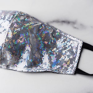 May include: A silver face mask with a holographic iridescent pattern. The mask has a black elastic band.