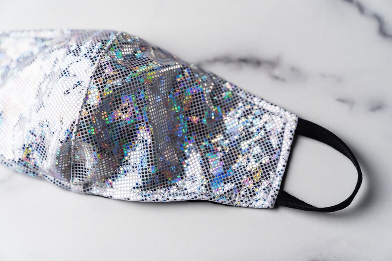 May include: A silver face mask with a holographic iridescent pattern. The mask has a black elastic band.