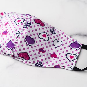 May include: A white face mask with a pattern of purple, pink, and white hearts and pink polka dots. The mask has black elastic ear loops and is made of fabric. The mask is designed to cover the nose and mouth.