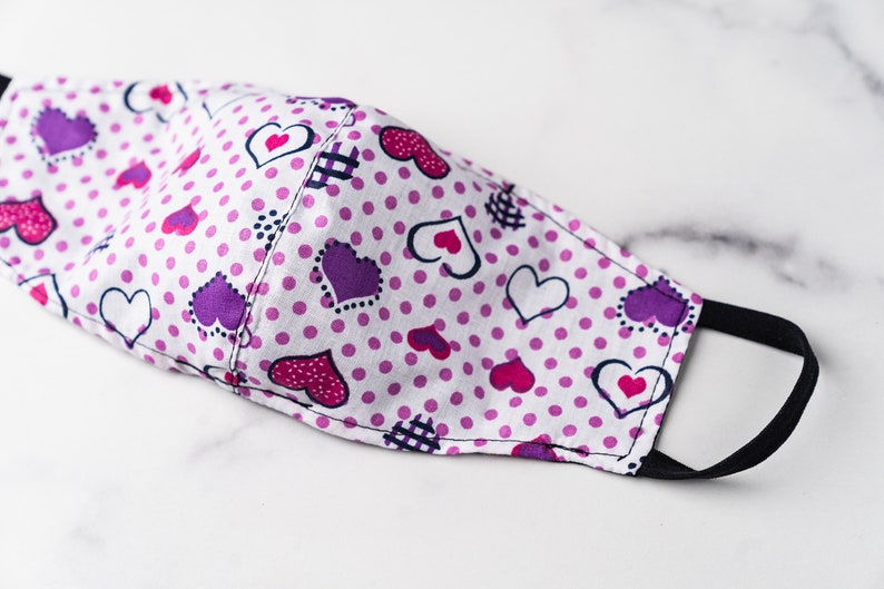 May include: A white face mask with a pattern of purple, pink, and white hearts and pink polka dots. The mask has black elastic ear loops and is made of fabric. The mask is designed to cover the nose and mouth.
