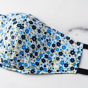 May include: A floral face mask with a white background. The mask features a pattern of small blue and navy flowers with green leaves. It has black elastic ear loops for secure wear.