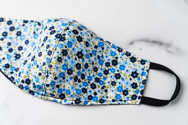 May include: A floral face mask with a white background. The mask features a pattern of small blue and navy flowers with green leaves. It has black elastic ear loops for secure wear.