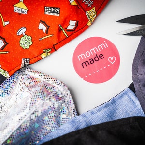 May include: A close-up of a variety of fabrics, including a red fabric with a school-themed pattern, a silver sequined fabric, a blue denim fabric, and a black fabric. The fabrics are arranged on a white surface with a pink circle sticker that says "mommi made" with a heart.